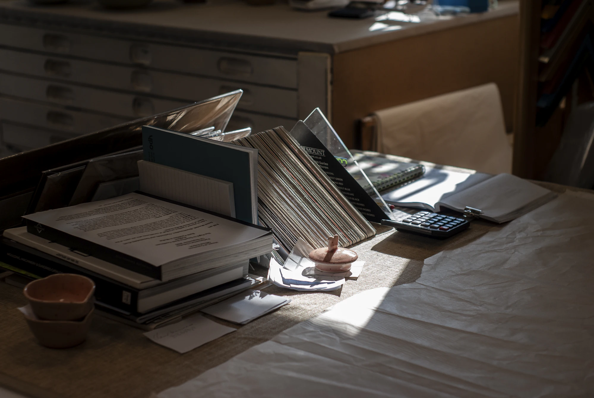 A sunlit desk with books, papers and an in-tray