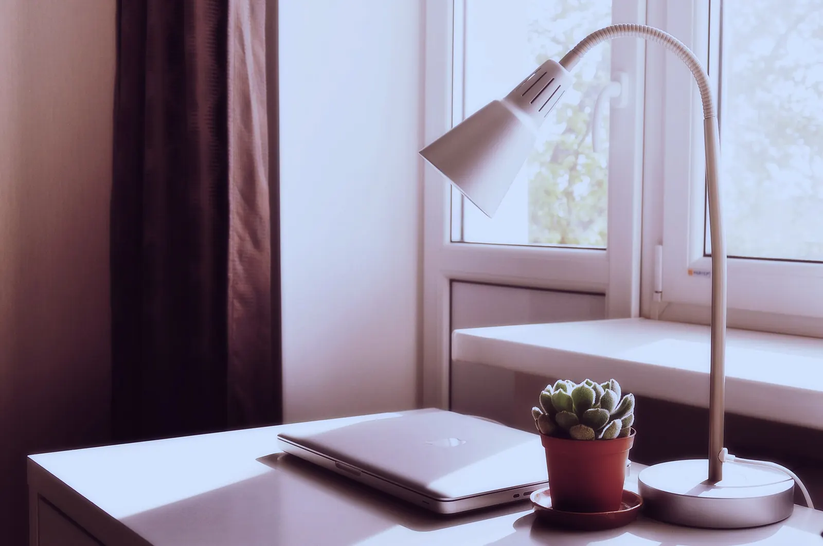 A quiet desk by a window with a lamp and a closed laptop